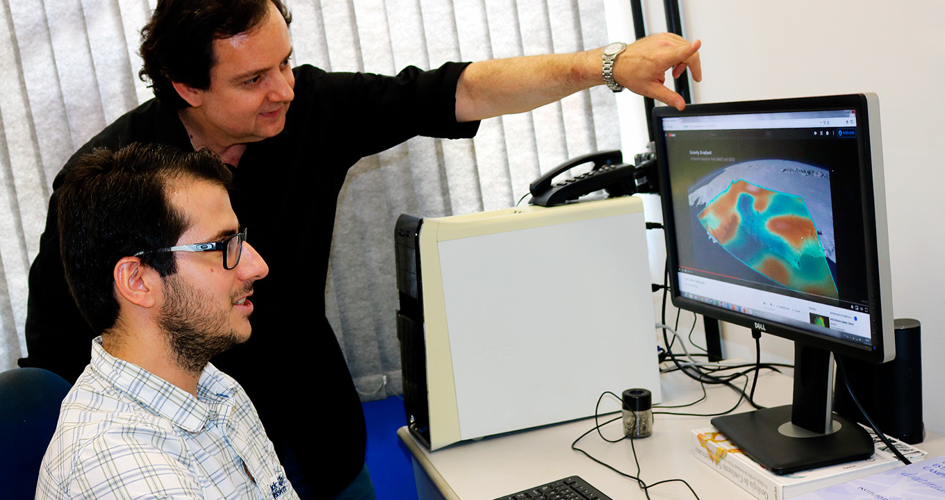 O professor Carlos Roberto de Souza Filho (em pé) e o doutorando João Motta | Foto: Divulgação Audiodescrição: em sala de trabalho, imagem em perspectiva, dois homens com olhares voltados para a direita da imagem, um ao lado do outro, sendo um sentado, em primeiro plano, e outro em pé, e inclinado para frente e com o braço esquerdo esticado para frente, com a mão tocando a parte superior de um monitor de lcd que está à frente deles em uma mesa. Ao lado do monitor, à esquerda, uma cpu branca. O homem sentado olha para o monitor à frente. Às costas deles, ao fundo uma persiana vertical. Imagem 1 de 1.