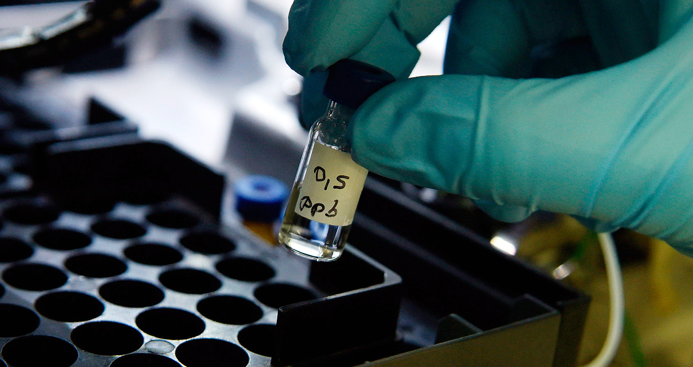 Amostras no Laboratório de Química Ambiental do Instituto de Química da Unicamp | Foto: Antonio Scarpinetti Audiodescrição: em área interna, imagem em perspectiva e close-up, frasco-amostra de vidro transparente cilíndrico, com cerca de seis centímetros de altura, contendo líquido transparente é depositado em um tipo de placa de plástico com dezenas de furos arredondados, onde o frasco será encaixado. O frasco é depositado por uma pessoa á direita na imagem, cuja foto mostra apenas as pontas dos dedos da mão direita, usando luva de silicone na cor verde. Há uma etiqueta no frasco, onde se lê 0,5ppb. Imagem 1 de 1
