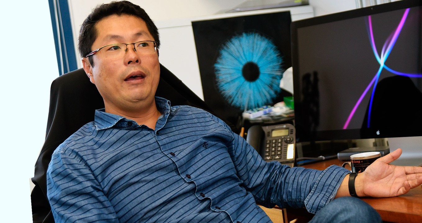 O professor Jun Takahashi, em sua sala no IFGW: admissão do Brasil no CERN conferiria prerrogativas importantes ao país | Foto: Antonio Scarpinetti Audiodescrição: em sala de trabalho, imagem em perspectiva e de busto, homem sentado em cadeira com olhar voltado para a direita da imagem, fala e mantém o braço direito abaixado e o esquerdo apoiado sobre uma mesa de madeira, à direita. Sobre a mesa há dois monitores de lcd, um ao lado do outro, que exibem imagens coloridas em fundo preto, e ainda caneca e telefone fixo. Há uma blusa preta apoiada no encosto da cadeira. Ele usa óculos e veste camisa de botões de mangas compridas em tom azul. Imagem 1 de 1.