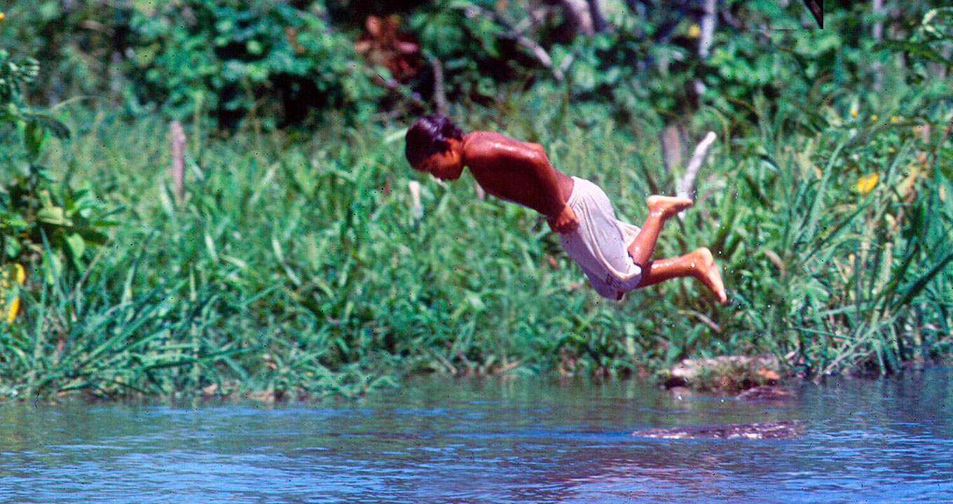 Criança mergulha no rio Solimões, no Amazonas: país tem a maior reserva mundial de água | Foto: José Sabino Audiodescrição: em área externa, imagem de corpo inteiro e em perfil, jovem indígena durante mergulho em rio. Ele está com o corpo na horizontal, durante o trajeto da queda, e mantém os braços junto ao corpo, lateralmente. Ele está com o corpo molhado e veste uma bermuda branca. Ao fundo, ampla área com densa vegetação. Imagem 1 de 1.