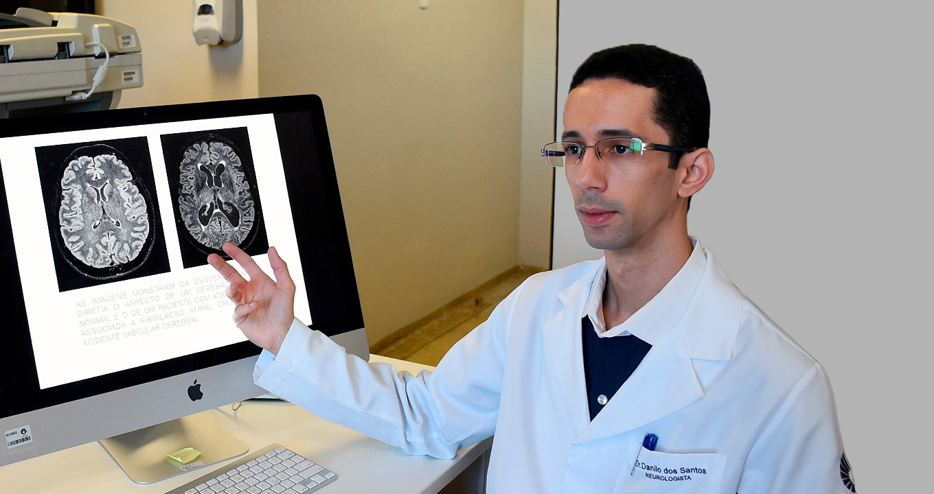 O médico neurologista Danilo Santos Silva, autor da tese: “Buscamos identificar precocemente pacientes suscetíveis ao desenvolvimento da demência, o que nos permitirá adotar medidas preventivas contra o avanço da doença” | Foto: Antoninho Perri Audiodescrição: em sala de exames de imagem, imagem em perspectiva e de busto, homem com olhar para a esquerda fala mantendo o braço direito projetado para o lado, apontando com dorso dos dedos para monitor de lcd com cerca de trinta polegadas. O monitor está sobre uma mesa retangular de madeira e exibe duas imagens em preto e branco de córtex cerebral. Atrás do monitor há uma parede com ampla janela de vidro transparente que permite ver partes de dois equipamentos. O homem usa jaleco branco. Imagem 1 de 1.