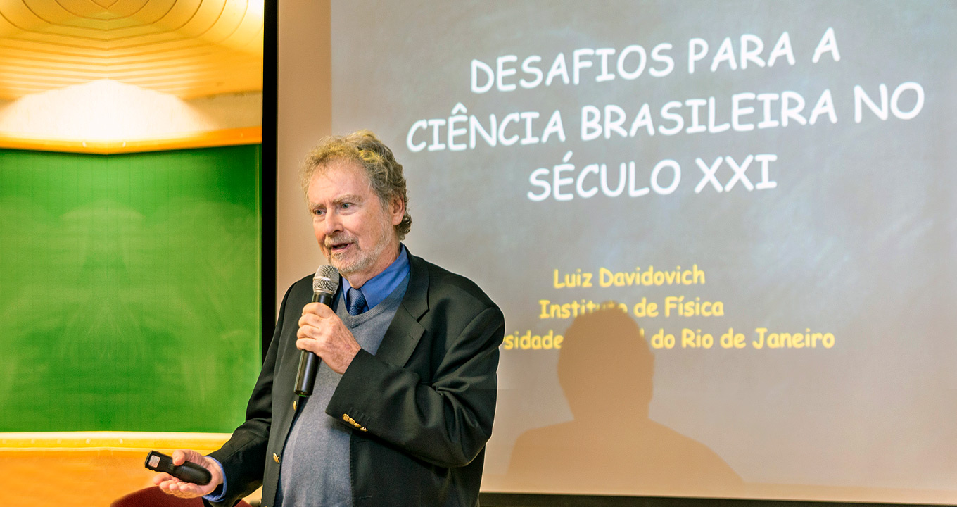 Luiz Davidovich, presidente da ABC, durante workshop Audiodescrição: em auditório, imagem de busto e em perspectiva, homem, em pé, fala em microfone sem fio segurando-o com a mão esquerda, mantendo o braço direito baixado e com um controle remoto na mão. Ele mantém o olhar voltado para a esquerda na imagem e veste terno cinza claro e gravata. Às costas, tela de projeção de data show onde se lê em destaque, em letras brancas, desafios para a ciência brasileira no século 21. Ao fundo, à esquerda na imagem, parte de uma lousa verde e luz no teto. Imagem 1 de 1.