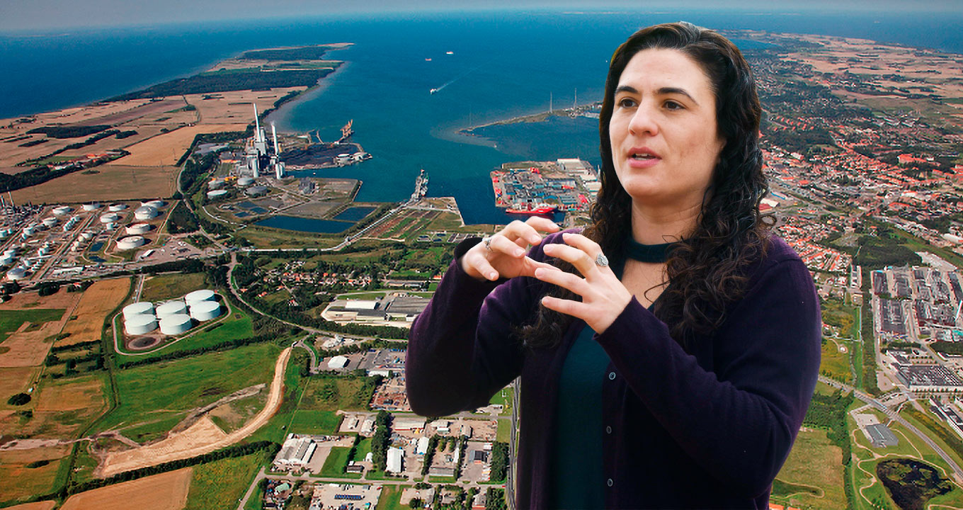O cluster de indústrias na cidade dinamarquesa de Kalundborg; no destaque, a arquiteta Thalita Dalbelo, autora de estudo e coordenadora do Plano Diretor Integrado da Unicamp Audiodescrição: montagem com duas imagens sobrepostas, sendo a primeira delas em perspectiva e de busto, de uma mulher em pé, à direita na imagem, e com olhar voltado para a esquerda. Ela fala e gesticula com as mãos, mantendo os braços dobrados à frente do corpo, com as mãos abertas quase encostadas uma na outra, como se segurasse uma esfera. Ela veste casaco roxo de lã. Na outra foto, ocupando todo fundo da imagem, tomada aérea de cluster de indústrias e mar avançando em parte do continente. Imagem 1 de 1