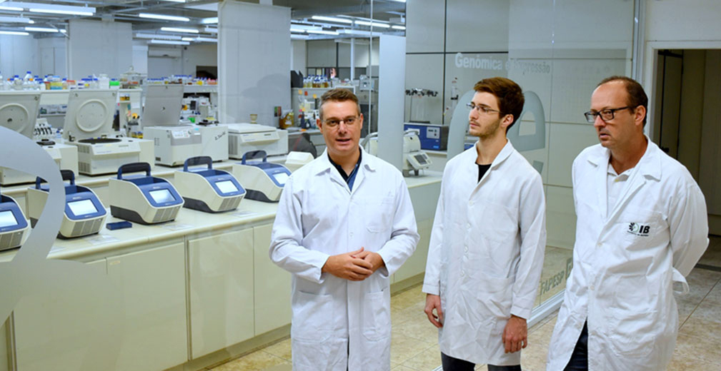 Orientador professor Fabio Papes (à esq.), pesquisador Antonio Pedro Camargo e coorientador Marcelo Falsarella Carazzolle | Foto: Antoninho Perri Audiodescrição: em laboratório, imagem frontal e plano médio, três homens em pé, um ao lado do outro, sendo que o primeiro à esquerda fala mantendo as mãos unidas à frente; o do centro observa-o com braços abaixados, e o outro mantém braços para trás. Todos usam óculos e jalecos brancos de mangas compridas. Às costas deles, à esquerda, imagem em perspectiva que se estende para o fundo da foto, ampla parede de vidro transparente, permitindo ver equipamentos do laboratório Genômica e Expressão. Imagem 1 de 1.