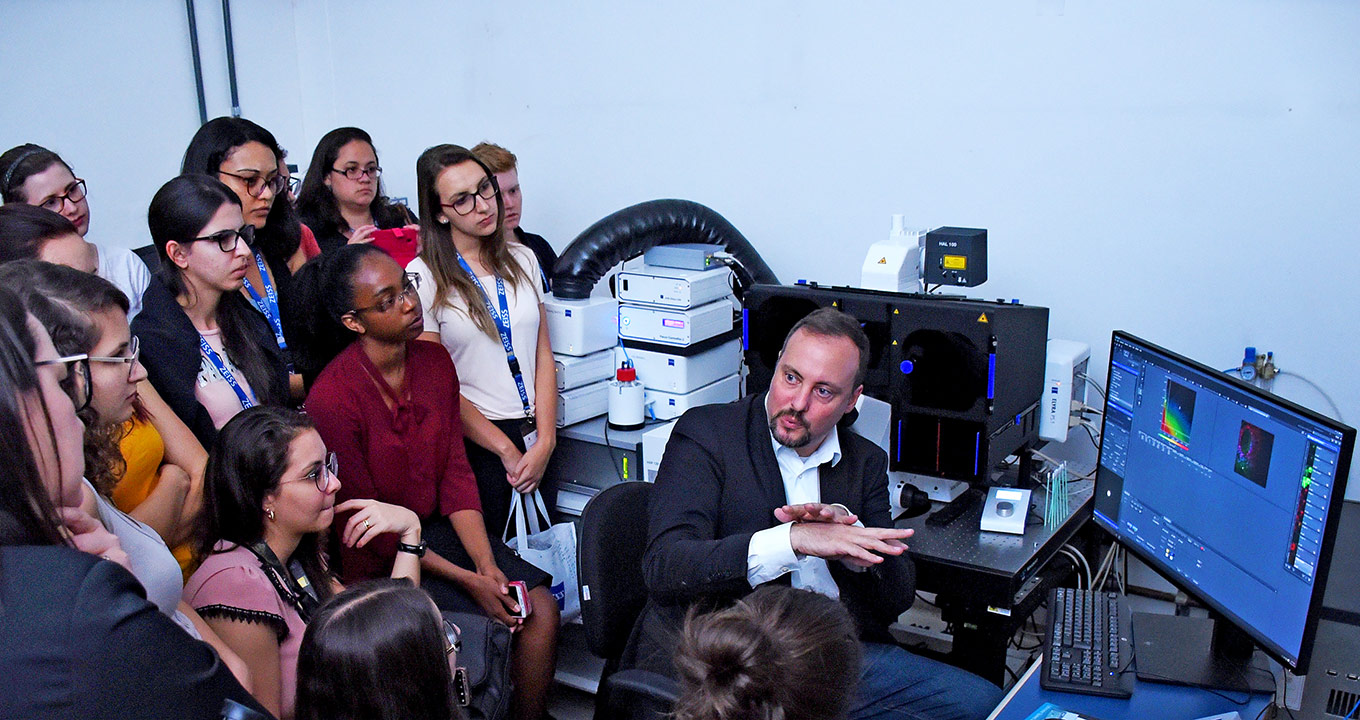 Christian Hellriegel, do Centro de Excelência Zeiss de Havard, explica como funciona o Super-Resolution | Foto: Antoninho Perri Audiodescrição: em uma sala, imagem frontal e de busto, homem sentado em cadeira fala e gesticula com as mãos mantendo braços dobrados e junto ao peito, com as palmas das mãos abertas, e a mão esquerda sobre a direita, encostadas. À frente dele e à esquerda na imagem, cerca de vinte jovens, alguns sentados e outros em pé, bem próximos uns dos outros, que o observam atentamente. Logo atrás do homem, uma mesa retangular de ferro com vários equipamentos, como caixas retangulares e monitor de lcd. Imagem 1 de 1