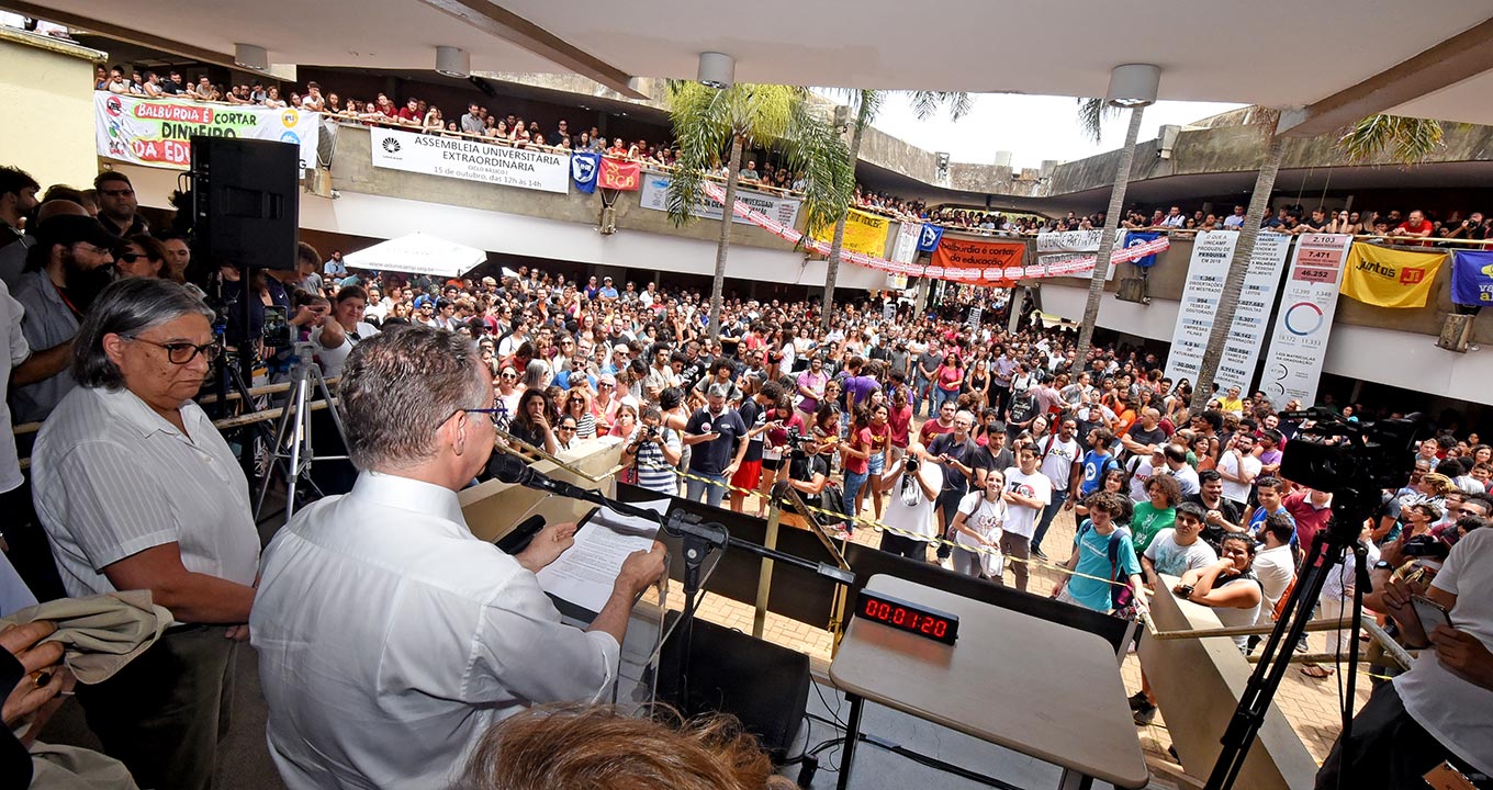 Audiodescrição: em área externa, imagem frontal e panorâmica centenas de pessoas em pé, aglomeradas uma ao lado da outra, em amplo pátio, participando de assembleia. Nas laterais, à direita e à esquerda e unidas ao centro, há duas edificações de três andares, com dezenas de pessoas aglomeradas nas sacadas. Há várias faixas verticais e horizontais afixadas ao longo das sacadas. Em primeiro plano, de costas, duas pessoas em pé, uma ao lado da outra. sendo que a da direita fala em microfone. Imagem 1 de 1.