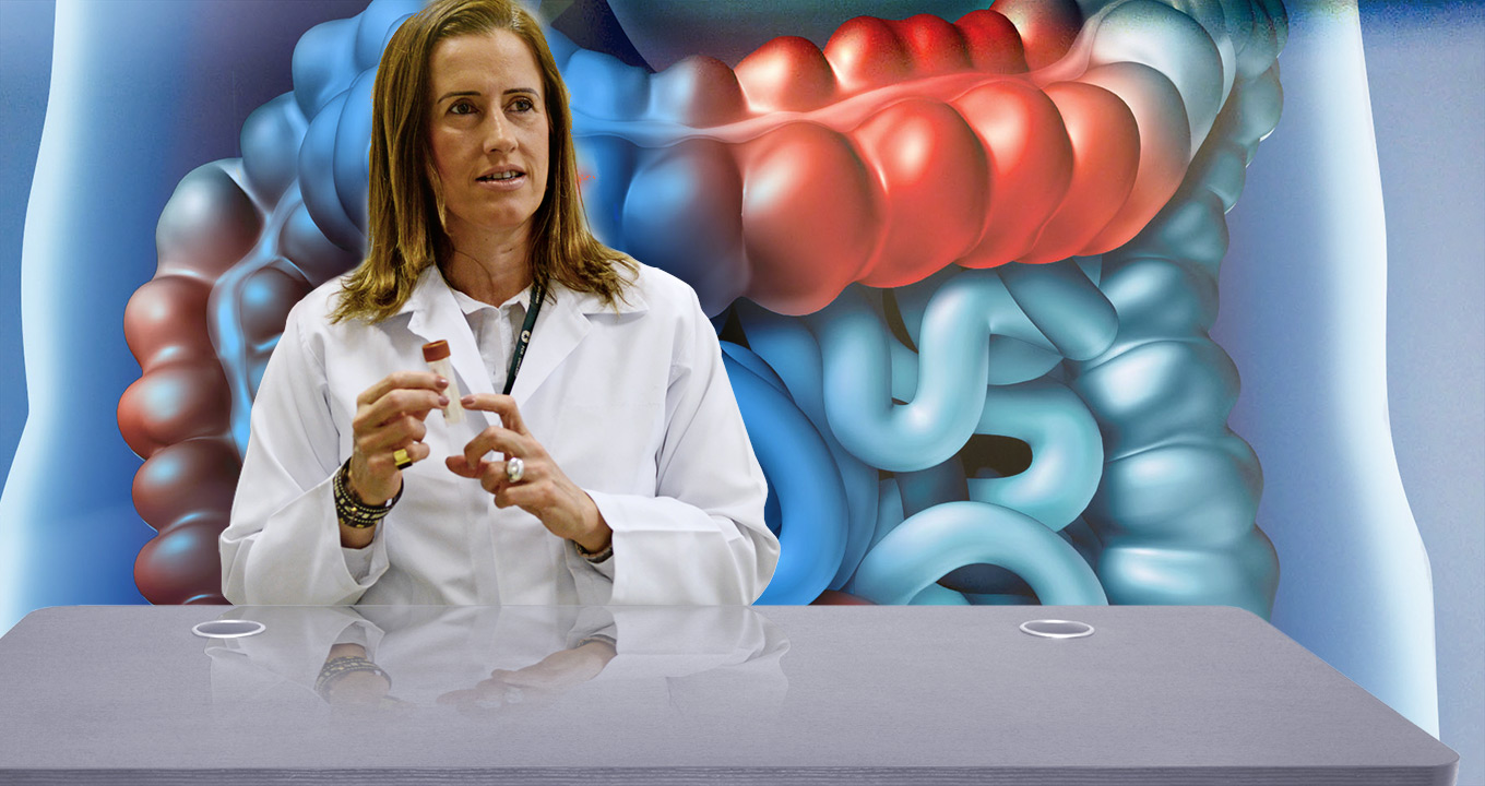 Daniéla Magro, nutricionista e pesquisadora do Departamento de Cirurgia da FCM Audiodescrição: em área interna, imagem frontal e de busto, mulher atrás da mesa, ao centro na imagem, fala e gesticula com as mãos mantendo os braços dobrados e à frente do peito. Ela olha sutilmente para a direita da imagem e usa jaleco branco de mangas compridas. Às costas dela, há uma ilustração que reproduz, em grande tamanho, um intestino humano, em tons de azul e vermelho. Imagem 1 de 1.