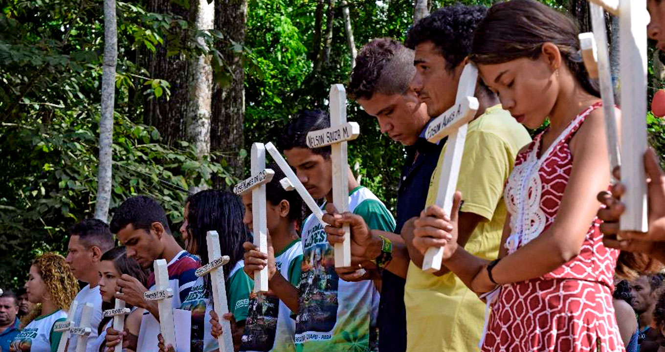 Audiodescrição: em área externa, imagem em perspectiva e de busto, grupo com cerca de 8 pessoas em pé, uma ao lado da outra, com olhar voltado para a esquerda da imagem. Todos seguram uma cruz de madeira pintada de branco, com cerca de 30 centímetros de largura por 50 de altura. Em cada cruz, há nomes escritos em preto, como Nelson Sousa e Edson Alves. Todos expressam tristeza, alguns com olhos fechados, outros cabisbaixo. Ao fundo, ao lado deles, área de vegetação alta e densa. Imagem 1 de 1.