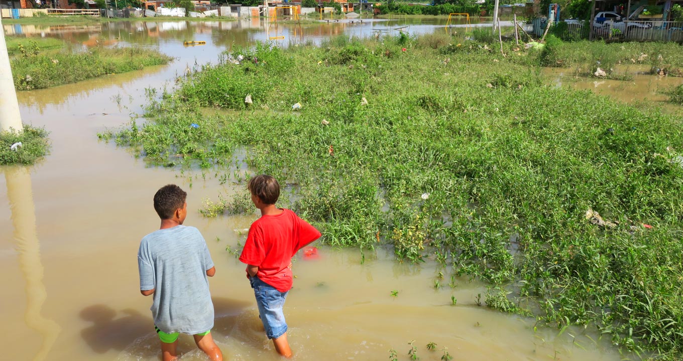 Audiodescrição: em área externa, imagem de corpo inteiro, dois jovens, de costas na imagem, à esquerda, caminham em ampla área alagada de um bairro. Eles vestem camisetas de mangas curtas, short e bermuda jeans. A água é turva e cercada por vegetação rasteira, à direita. Ao fundo da imagem, há um parquinho infantil alagado, com brinquedos como balanço e gangorra, fachadas de casas com frentes feitas de madeira e tijolos, e também um carro estacionado, à direita. O dia está ensolarado. Imagem 1 de 1.