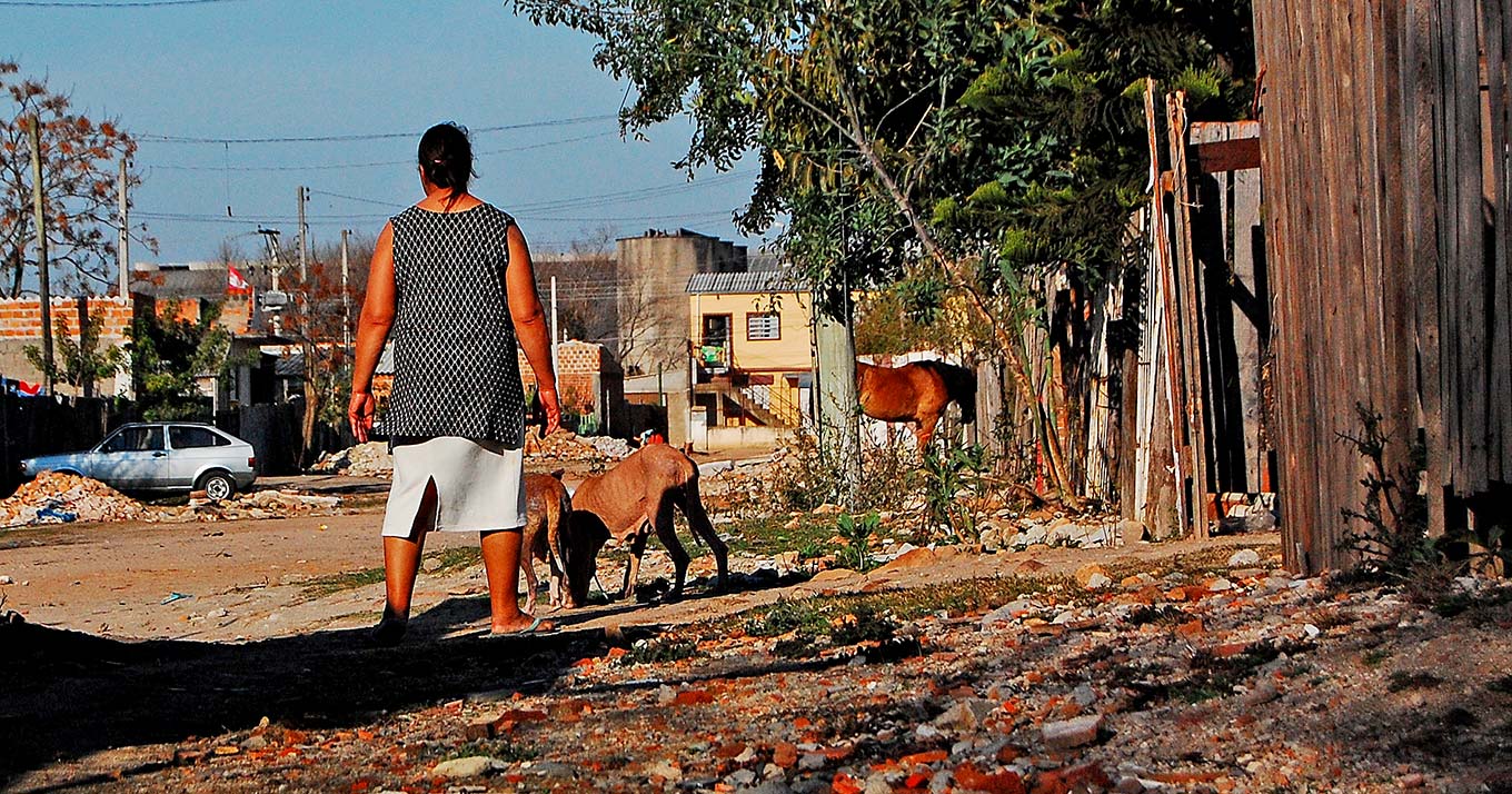 Medidas do governo federal colocam em xeque a sobrevivência dos serviços públicos e da população audiodescrição; fotografia colorida mostra uma senhora de costa observando a rua; ela está em uma rua de terra, com casebres de madeira em volta e cachorros estão ao seu lado