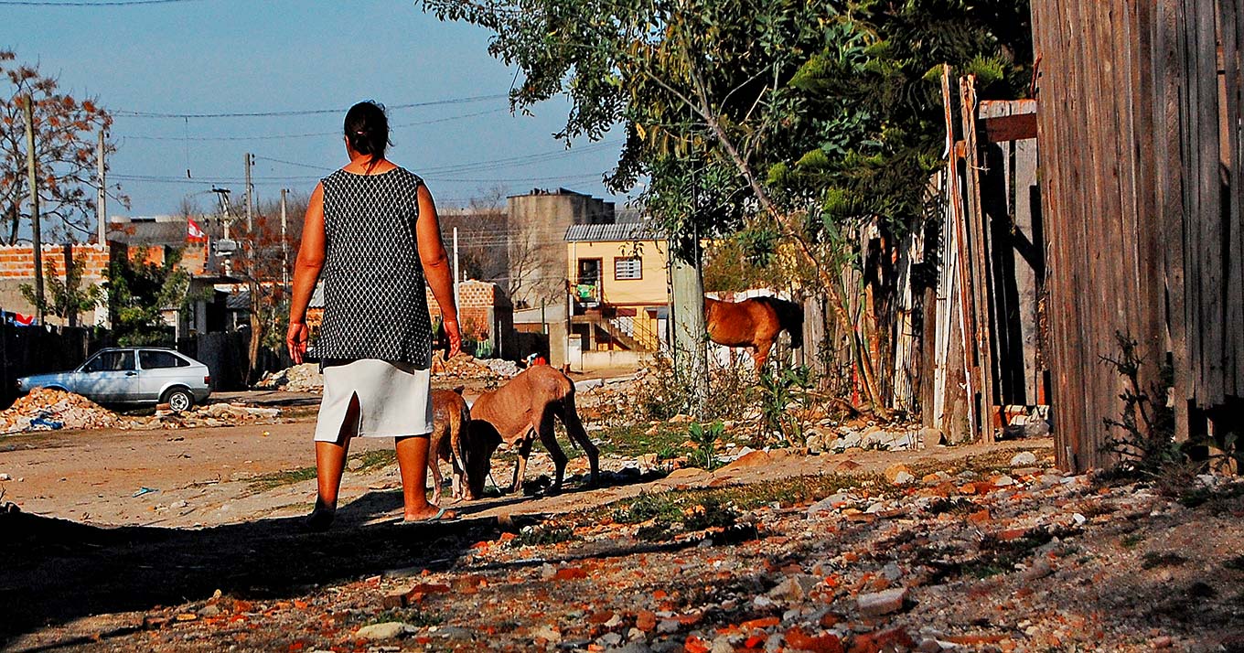 audiodescrição; fotografia colorida mostra uma senhora de costa observando a rua; ela está em uma rua de terra, com casebres de madeira em volta e cachorros estão ao seu lado