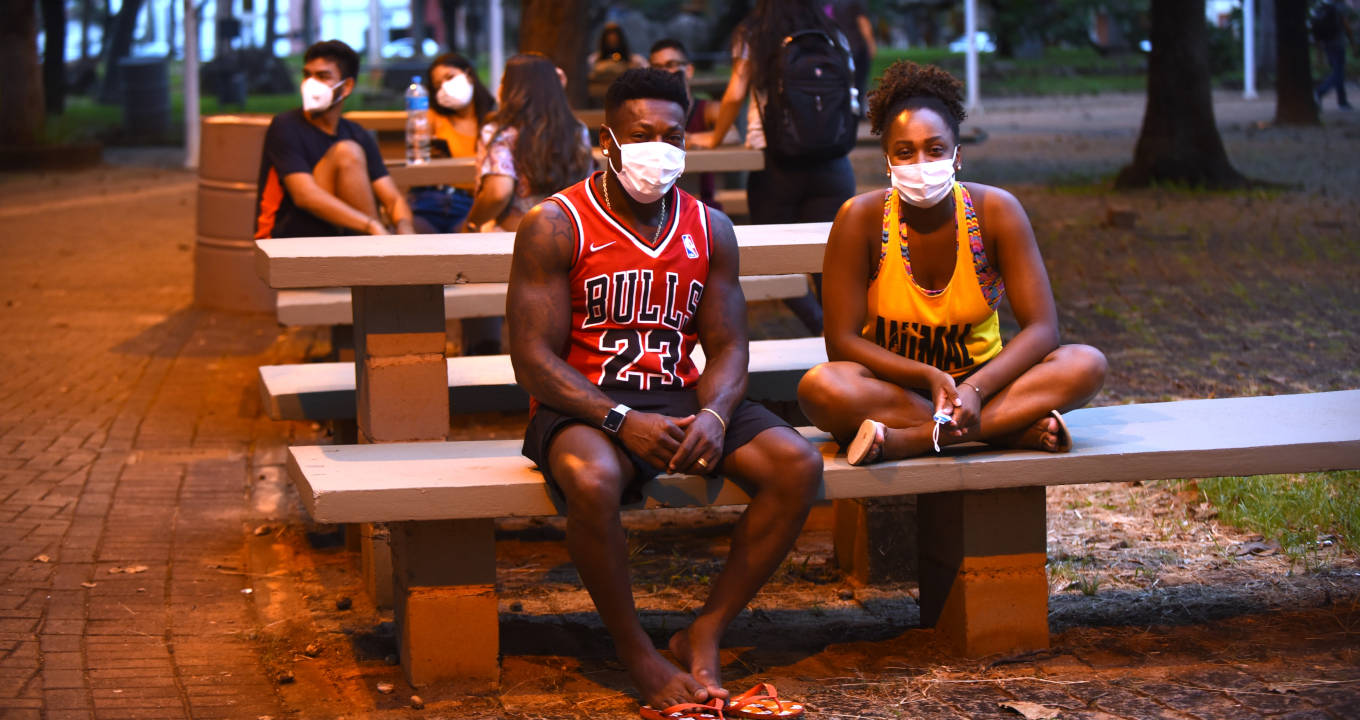 Lei de cotas alterou o perfil demográfico da população acadêmica da Unicamp Audiodescrição: Em uma praça ao anoitecer, imagem frontal e à curta distância de 2 pessoas sentadas em banco de concreto, sendo um homem, à esquerda da imagem, que mantém os braços apoiados sobre as pernas, com mãos unidas à frente, e uma mulher com as pernas cruzadas sobre o banco, e braços apoiados sobre as pernas, com mãos unidas à frente. Atrás deles há outros bancos e mesas de concreto com pessoas sentadas. Ambos usam máscara de proteção respiratória e camiseta regata. Imagem 1 de 1.