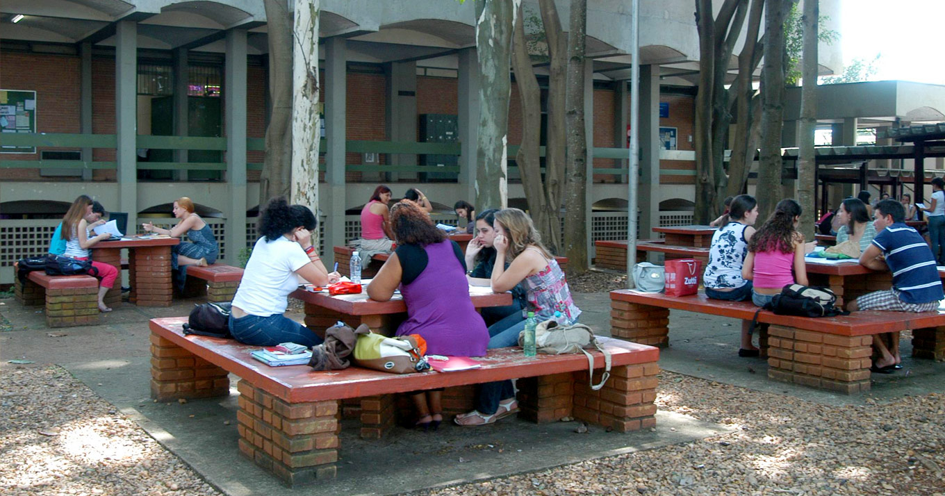 Curso de Estudos Literários lança a editora-escola Asa da Palavra Audiodescrição (AD): em área externa, imagem em plano geral de 4 grupos com cerca de 4 a 5 pessoas sentadas em bancos de concreto ao redor de várias mesas retangulares de concreto distribuídas no local. Elas apoiam braços nas mesas, sendo que algumas pessoas estudam e outras conversam. Há bolsas e mochilas nos bancos. O piso no local onde apoiam os pés é de concreto, havendo, no entorno, pedras de rio decorativas. Há altas árvores que fazem sombra no local. Ao fundo, edificação de concreto. Imagem 1 de 1.