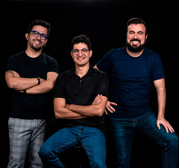 Imagem mostra três homens de pé. Os três são brancos, usam camiseta preta e estão sorrindo.