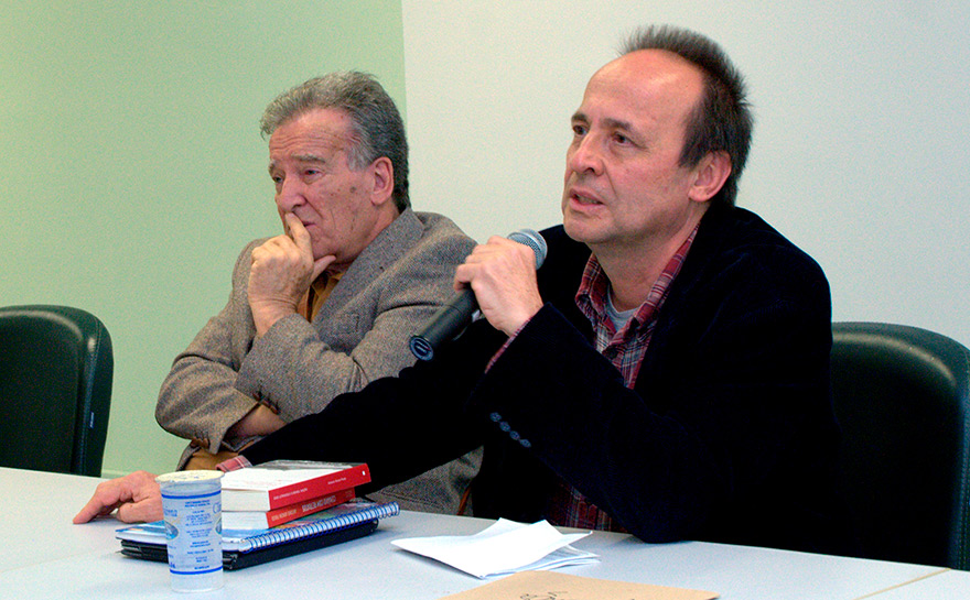 Foto de dois homens sentados em um mesa. O homem à esquerda é branco, tem cabelo grisalho, está usando um blazer e está com os braços cruzados, apoiando a cabeça em uma das mãos.