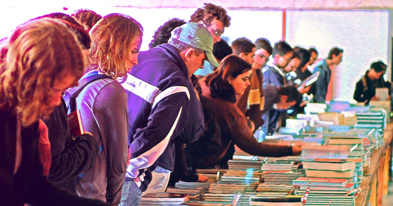 Foto de um grupo de pessoas de pé em um ambiente aberto. Elas estão escolhendo livros espalhados em mesas.