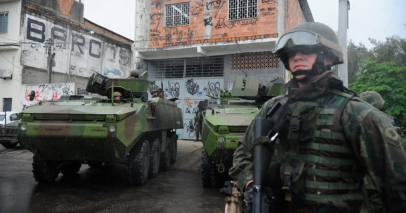 Foto que mostra um soldado em primeiro plano, ele usa capacete, óculos e está segurando um rifle, à esquerda há um tanque e ao fundo um casa de dois andares com tijolos aparentes.