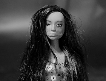 Foto em preto e branco que mostra a cabeça de uma boneca. Ela tem cabelos compridos e lisos e tem um curativo em um dos olhos.