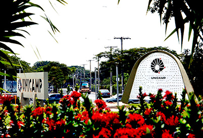 Imagem colorida, no lado esquerdo da imagem está uma placa de concreto com o logo e o nome unicamp, do lado diretiro está o um arco de concreto com uma lona branca e o logo da Unicamp impresso ao centro